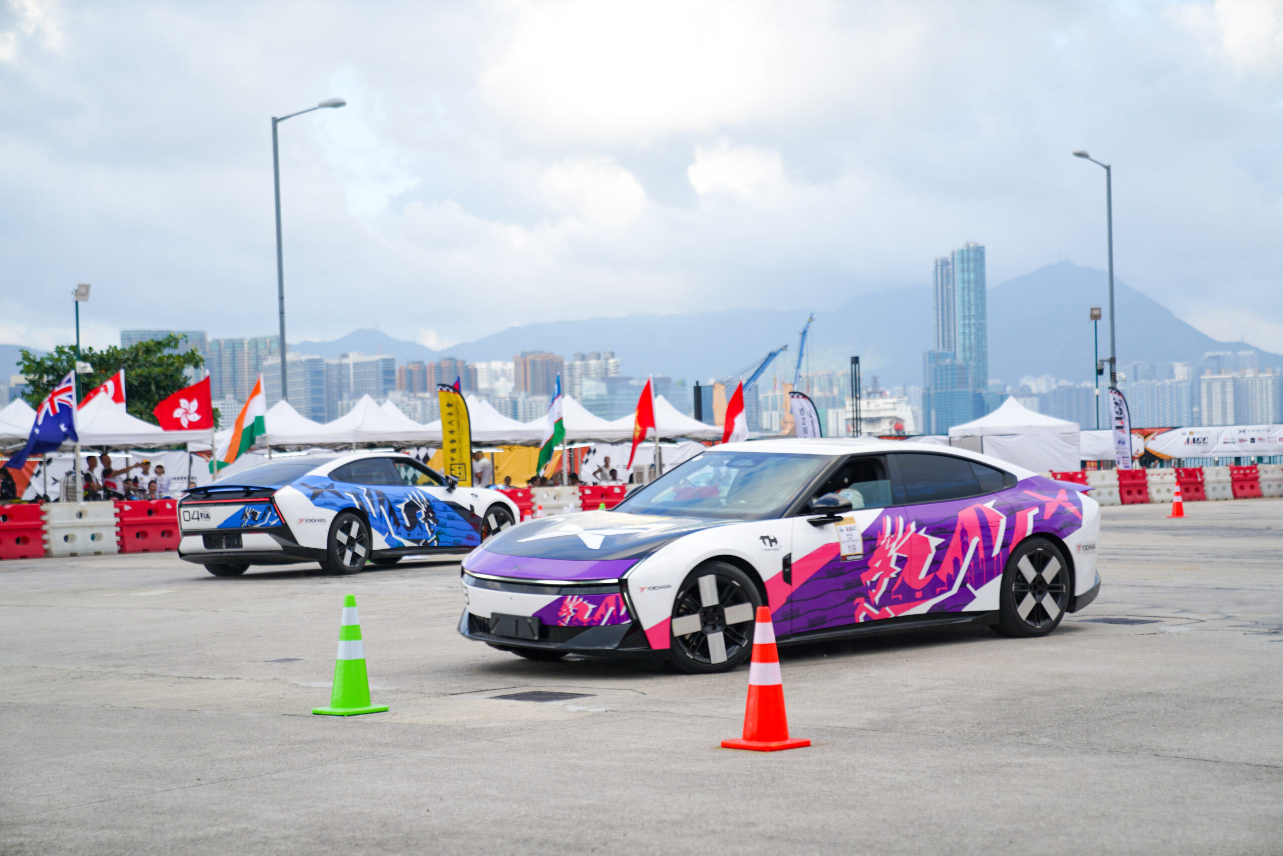 Two XPENG New P7s with race numbers 19 and 04 on the cone course on the Hong Kong waterfront, with flags and tents behind