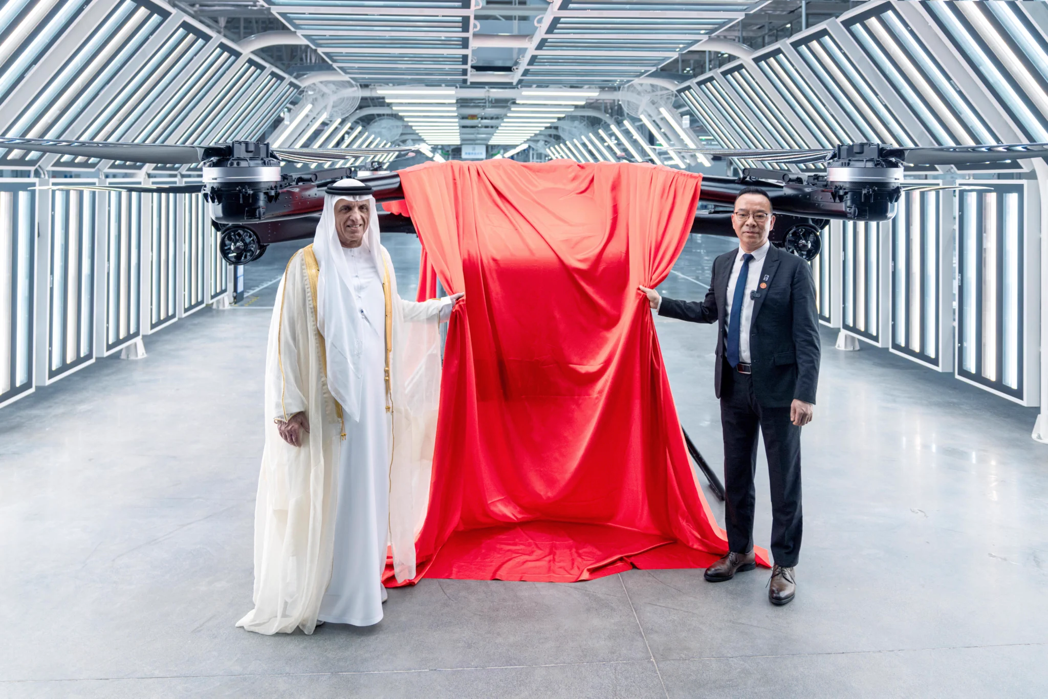 Two men standing either side of a large red cloth being drawn back to reveal a flying car with rotors in a lit hall