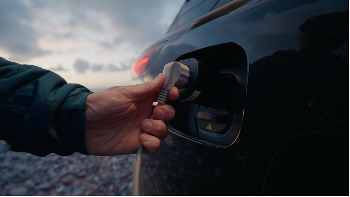 A hand pushing the white V2L adapter into the charge port of a dark XPENG at dusk