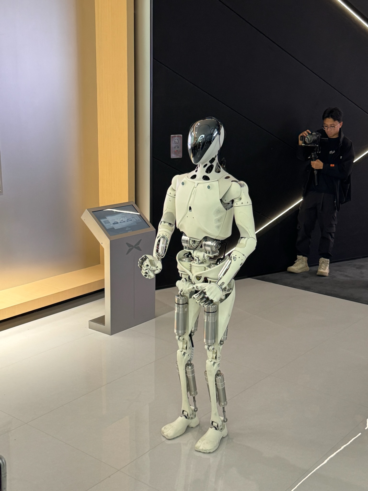 White IRON robot with exposed waist and leg mechanisms beside an exhibition information screen