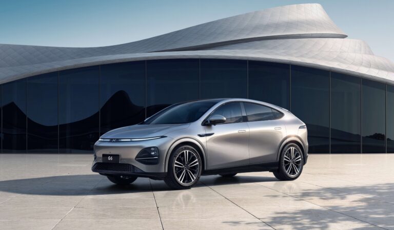 Silver XPENG G6 parked on a stone plaza in front of a white curved-roof building, front three-quarter view