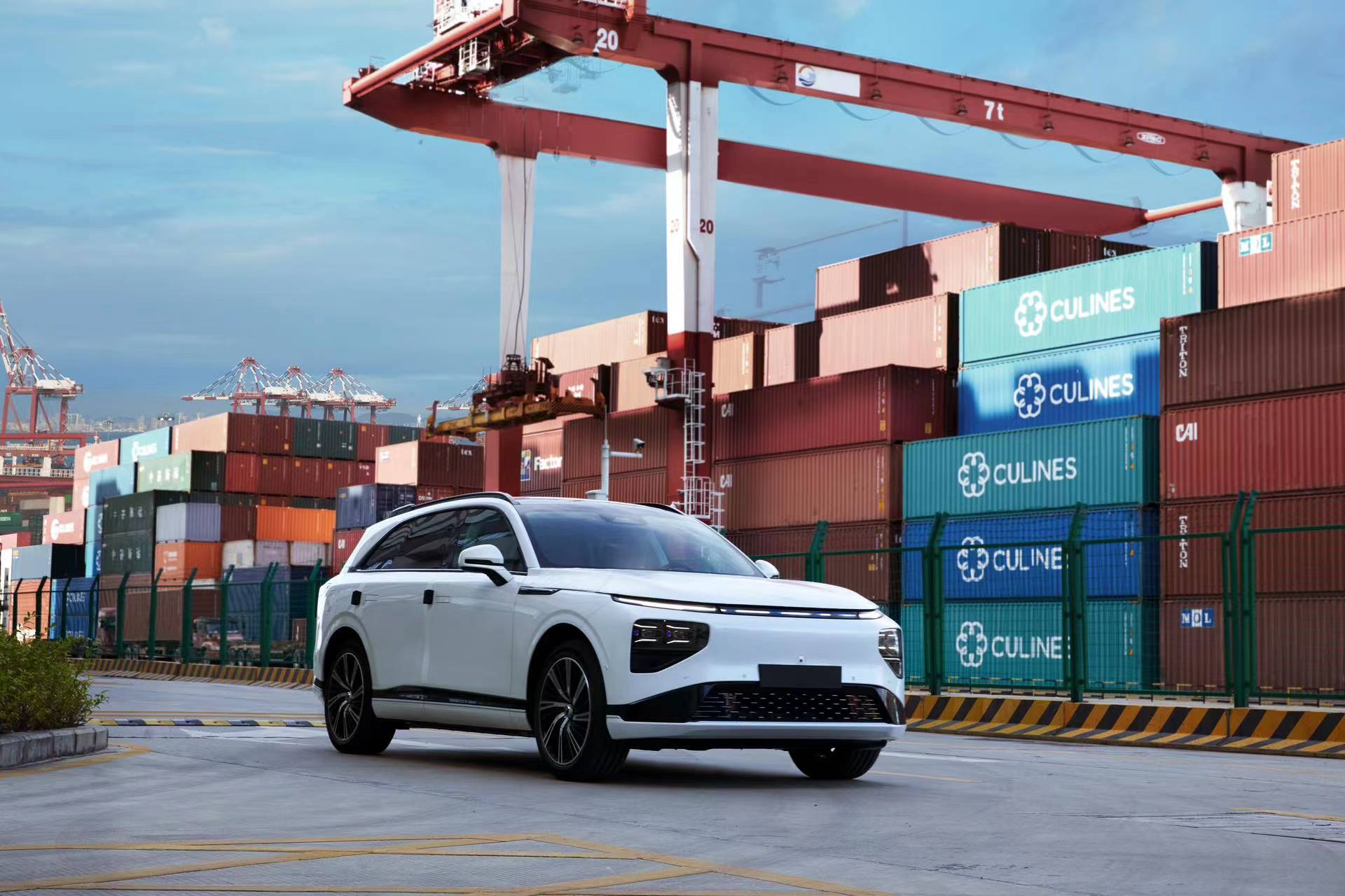 White XPENG G9 on a container terminal apron with stacked containers and a red gantry crane behind