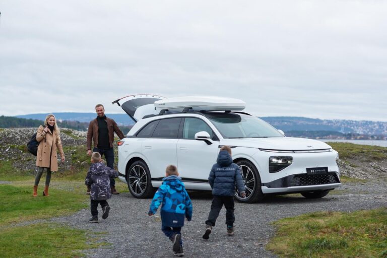 White XPENG G9 with the tailgate up and a roof box fitted on a gravel headland by the Oslofjord, a family walking towards it
