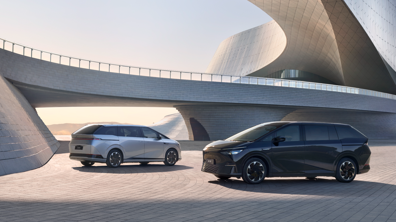 Two XPENG X9 MPVs, one silver and one black, on a paved plaza in front of a sweeping concrete landmark at dusk