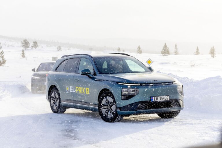 Teal grey XPENG G9 with yellow EL-PRIX decals driving on packed snow in falling snow, a second test car behind