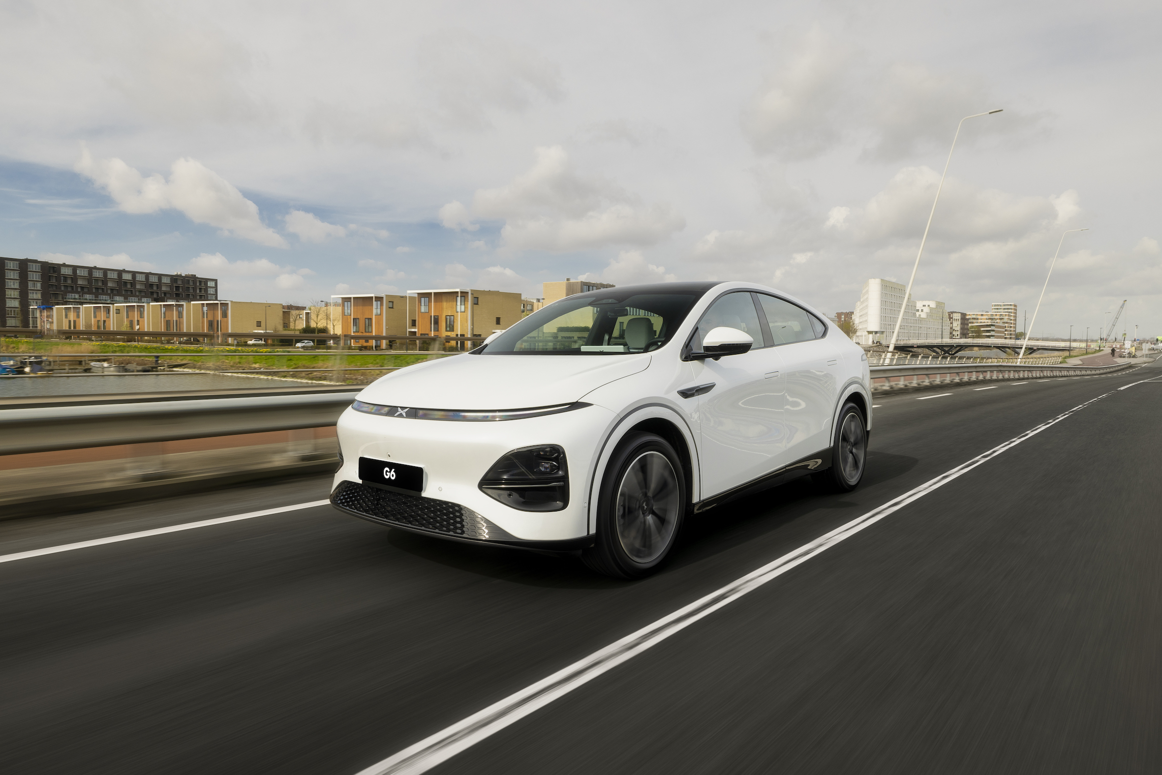 Hvid XPENG G6 i fart på en hollandsk motortrafikvej forbi boliger ved vandet i lavt sollys