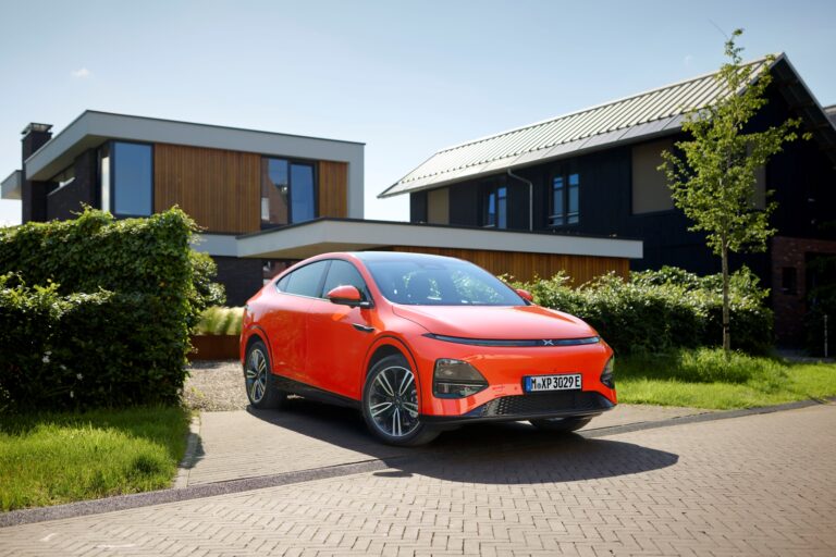 Orange XPENG G6 parked on a block-paved drive in front of a modern dark-timber house