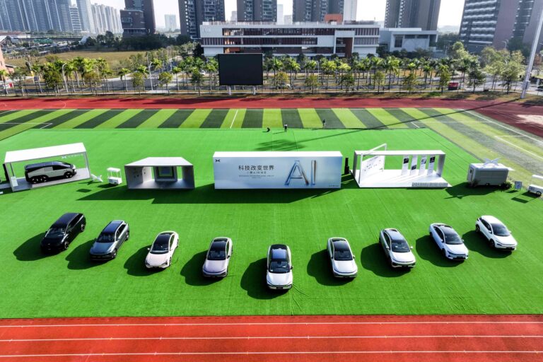 Nine XPENG cars lined up on a green pitch inside a running track, with event pavilions and an AI Day banner behind