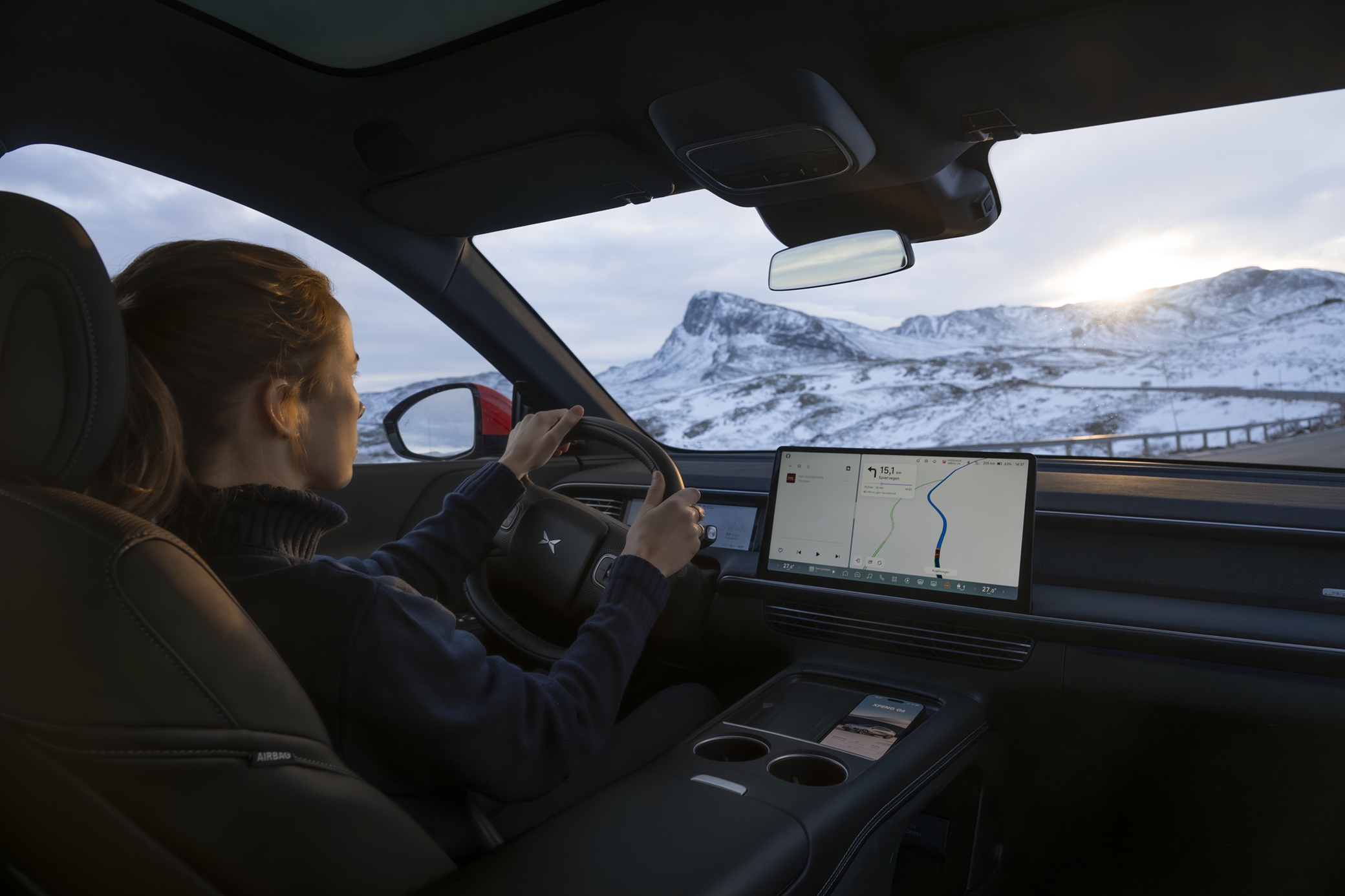 View over a driver's shoulder in an XPENG G9 on a snow-covered mountain road, the centre screen showing navigation