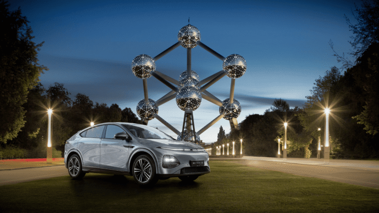 Silver XPENG G6 parked on grass at blue hour with the Brussels Atomium lit behind it