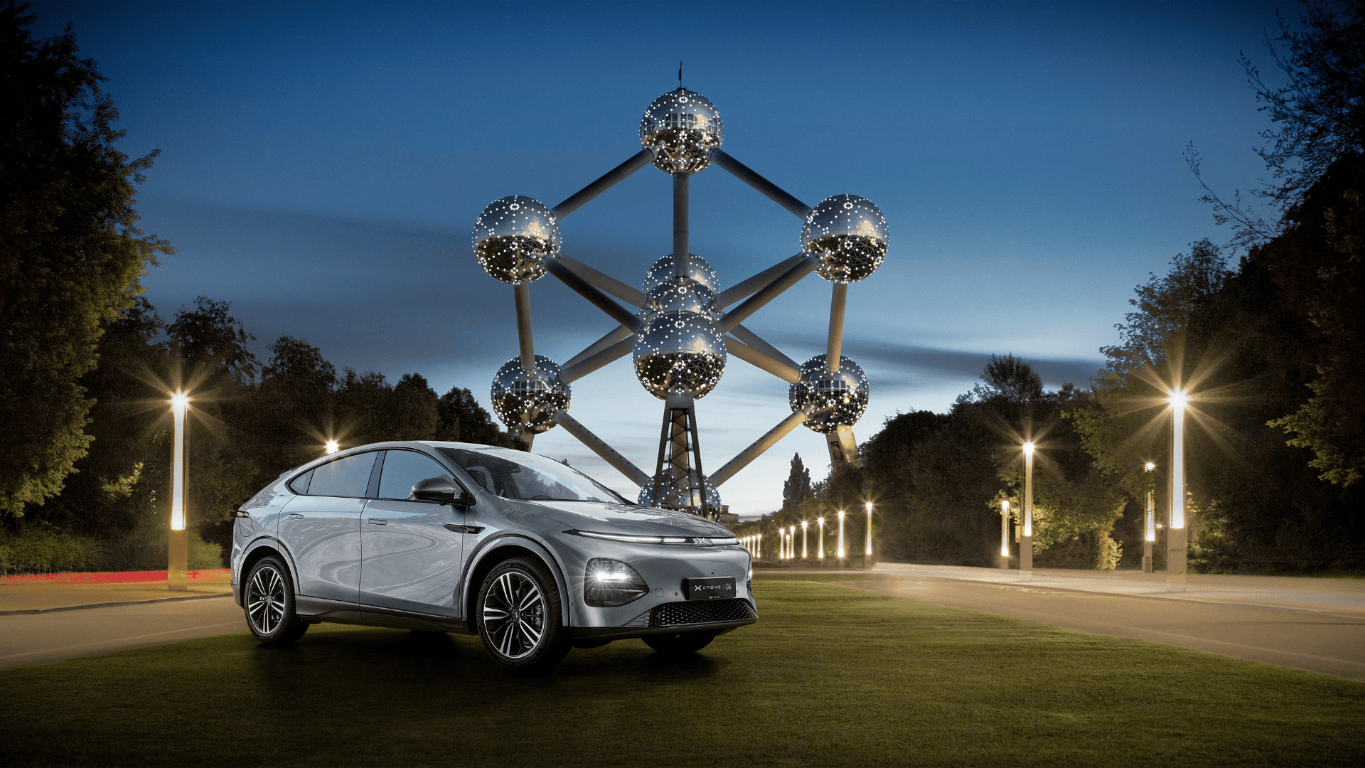 Silver XPENG G6 parked on grass at blue hour with the Brussels Atomium lit behind it