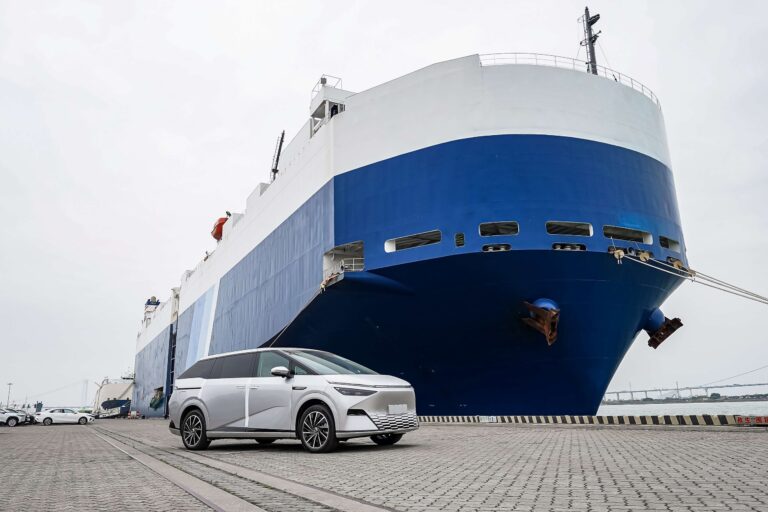 White XPENG X9 on a quayside in front of the blue bow of a car carrier