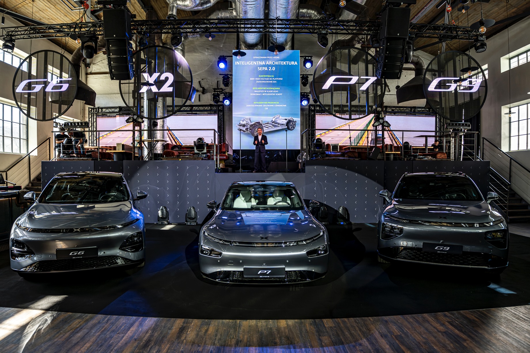 Three silver XPENG cars on a launch stage under illuminated ring letters, a speaker on the podium behind