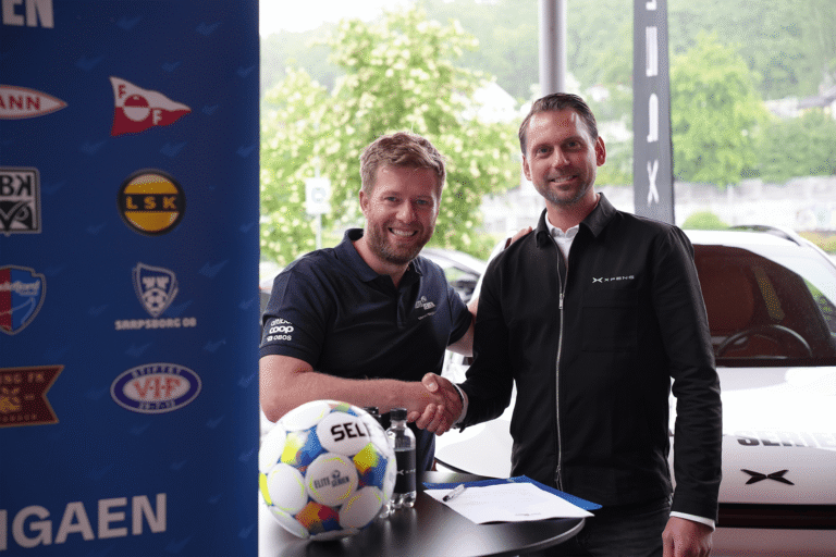 Two men shaking hands over a signed contract with an Eliteserien ball on the table and a white XPENG behind them