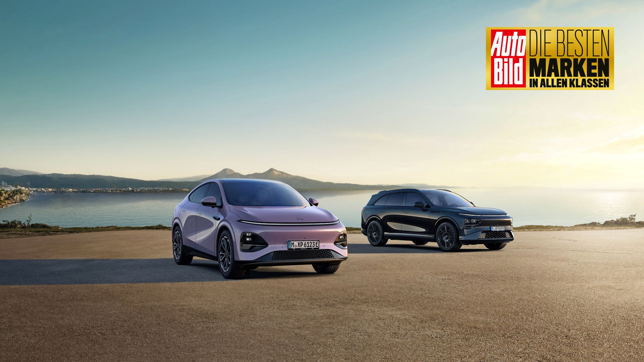 Pale lilac New XPENG G6 and a black New G9 on a coastal apron at sunset, with an Auto Bild award badge in the corner
