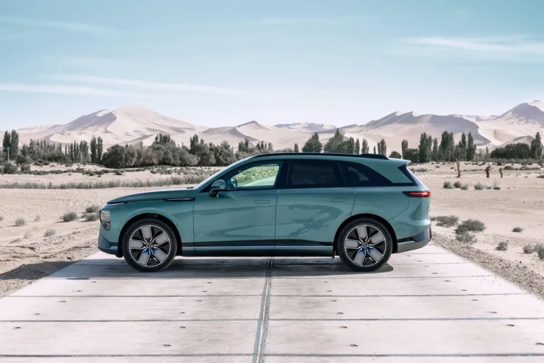 Green XPENG G9 seen from the side on a concrete strip in the desert, with sand dunes and a row of trees behind it