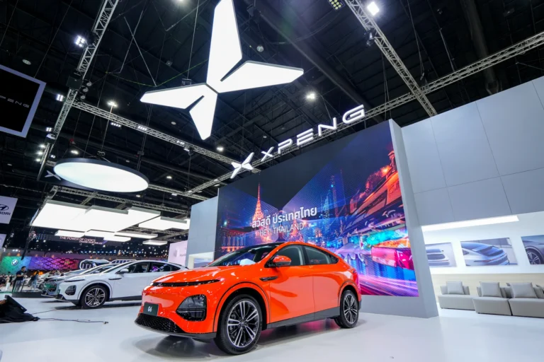 Orange XPENG G6 on XPENG's stand at the Bangkok Motor Show, in front of a screen reading Hey! Thailand, with the XPENG logo above the stand