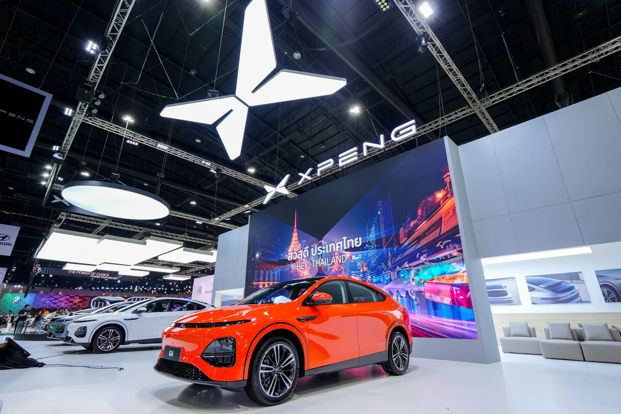 Orange XPENG G6 på XPENGs stand på Bangkok Motor Show foran en skærm med teksten Hey! Thailand og XPENG-logoet over standen