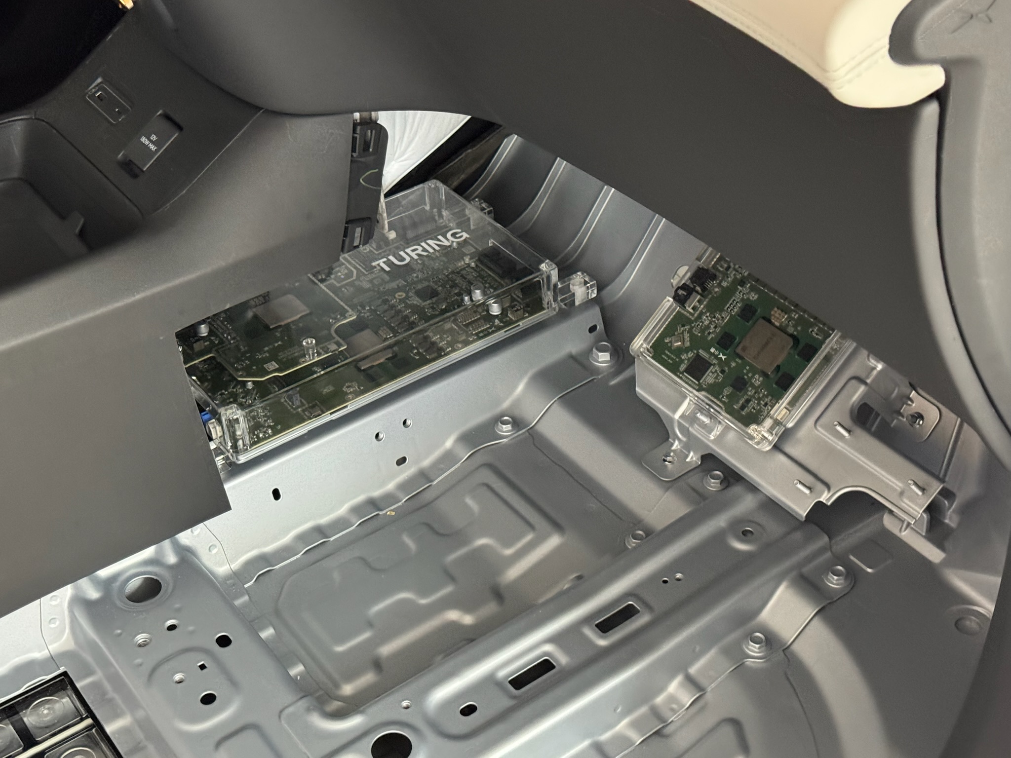 The Turing driving computer in a cutaway XPENG: a circuit board under a clear cover marked TURING, bolted to the floor pan beside a smaller module